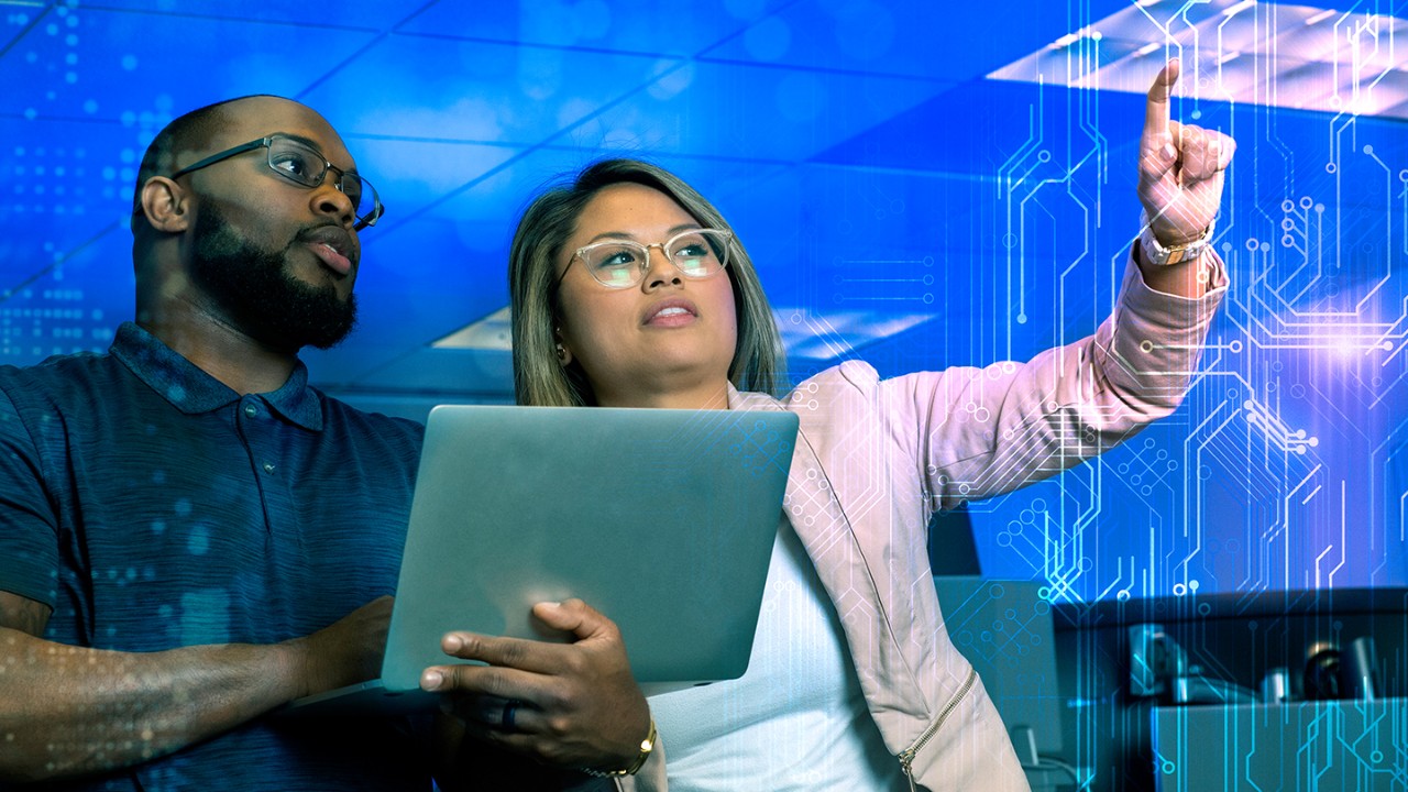 Man standing and holding a laptop with a woman standing next to him, pointing at data connections on a touch screen.