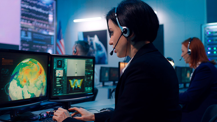 Woman with a headset on looking at dual computer screens.